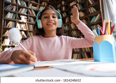 Happy indian latin preteen girl school pupil wearing headphones raising hand distance learning online at virtual lesson class with teacher tutor by video conference call at home, webcam view.