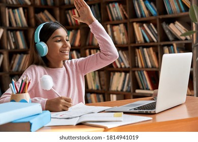 Happy indian latin preteen girl school pupil wearing headphones raising hand distance learning online at virtual lesson class with teacher tutor on laptop by zoom video conference call at home.