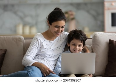 Happy Indian little son and loving mom enjoying leisure at laptop on couch, watching movie, using app, chatting online. Mother and child boy shopping on internet at home, talking on video call