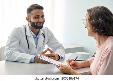 Happy Indian male doctor consulting senior old patient filling form at consultation. Friendly professional physician talking to mature woman signing medical paper during appointment visit in clinic. - Powered by Shutterstock