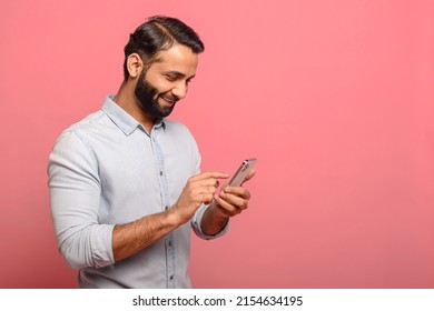 Happy Indian man in casual jeans shirt using smartphone isolated on pink, hispanic guy chatting online, using new mobile app, texting with girlfriend
