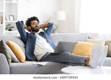 Happy indian man in casual wear reclining on sofa with his laptop, cheerful eastern guy resting on couch at home with hands behind head, looking satisfied, taking break from remote work - Powered by Shutterstock