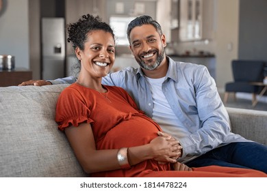 Happy Indian Man Embracing His Pregnant Wife And Looking At Camera. Loving Husband Sitting With Pregnant African American Wife Hand On Stomach. Portrait Of Excited Mid Adult Couple Expecting A Baby.