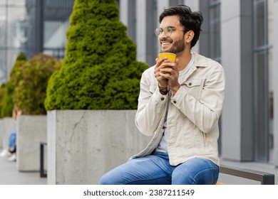 Happy Indian man enjoying morning coffee hot drink and smiling outdoors. Relaxing, taking a break. Arabian guy in city downtown street sitting on bench, drinking coffee to go. Town lifestyles outside