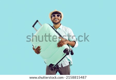 Happy Indian man going on holiday trip. Studio shot of cheerful handsome South Asian traveler in panama hat and stylish sun glasses leaving for vacation abroad, holding his mint suitcase and smiling