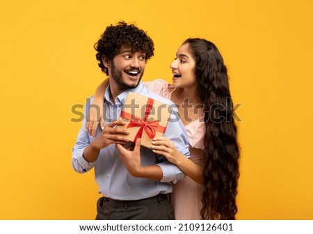 Happy indian man got present from his loving girlfriend, posing with gift box and smiling to each other while his woman cuddling him from back, standing over yellow background