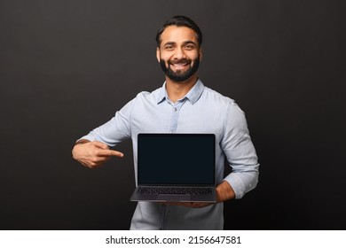 Happy Indian Man Holding Laptop With Empty Screen And Points Finger At It, Hispanic Male Employee Presenting New Computer App, Promotion And Advertise Concept