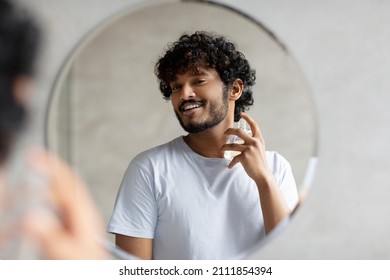 Happy Indian Man Looking In Mirror Spraying On Cologne In Bathroom Interior, Doing Morning Skincare Routine. Beauty Skin Care, Moisturizers Products And Perfume