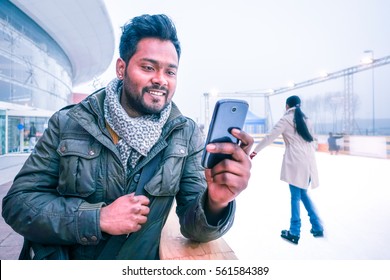 Happy Indian Man Looking Mobile Phone Outdoors At Winter With Ice Skating Scene Background - Cheerful Bangladeshi Young Guy Using Cellphone Smiling Looking Telephone Screen Outside On Grey Light Day