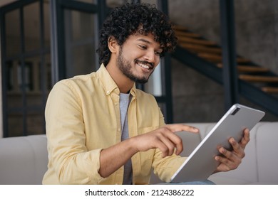 Happy Indian Man Shopping Online, Ordering Food, Reading Good News Sitting On Sofa. Smiling Asian Freelancer Using Digital Tablet, Receive Payment Working Online From Home