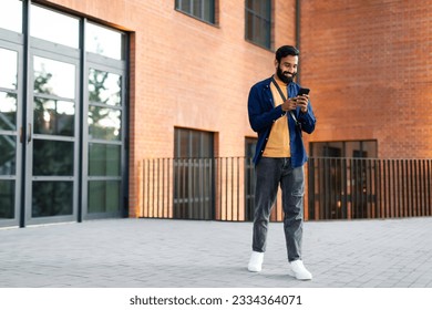 Happy indian man texting on mobile phone using great application standing near residential building in urban area outdoor. Guy with smartphone reading messages, full length, copy space - Powered by Shutterstock