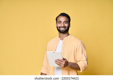 Happy Indian Man Using Digital Tablet Standing Isolated On Yellow Background. Smiling Ethnic Guy Holding Pad Wearing Earbud Advertising Virtual Class Webinars Or Audio Books.