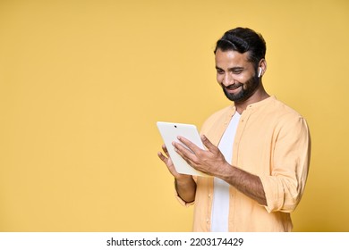 Happy Indian Man Using Digital Tablet Standing Isolated On Yellow Background. Smiling Ethnic Guy Holding Pad Wearing Earbud Watching Virtual Class Webinar, Having Remote Video Call.