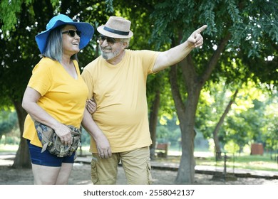 Happy indian mature couple wearing hat having eyewear sunglasses at park. Senior people enjoying retirement life. Senior Couple Enjoying Picnic Together having fun and pointing fingers aside way - Powered by Shutterstock