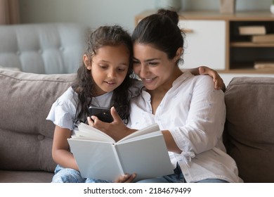 Happy Indian mom hugging little kid on couch, showing smartphone screen to girl on couch, using online app, browsing Internet. Mum and daughter reading paper book at home - Powered by Shutterstock