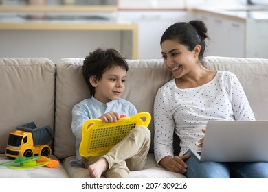 Happy Indian mom and little son enjoying leisure together, using toy drawing pad board and laptop, talking, smiling, laughing. Mother and kid watching movie, playing on couch at home