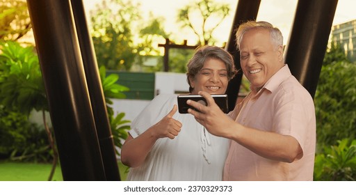 Happy Indian old retired senior couple standing together hold mobile phone taking funny faces selfie outdoor house. Smiling aged husband wife looking camera enjoying summer vacation outside home park - Powered by Shutterstock