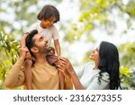 Happy indian parents playing with daughter while kid on father shoulder at outdoor - concept of parenthood, family bonding and weekend holidays