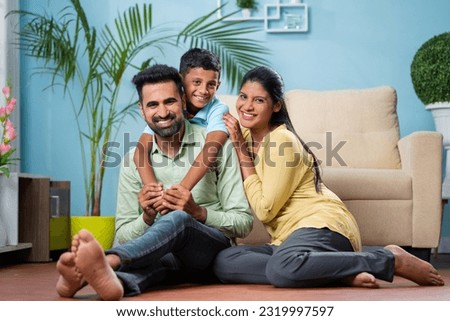 Happy indian parents with son by looking at camera while sitting on floor at home - concept of family bonding, weekend holiday and parenthood.