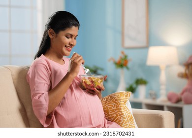 Happy indian pregnant woman eating fruit salad while sitting on sofa at home - concept of Pregnancy nutrition, Healthy eating and Prenatal wellness. - Powered by Shutterstock