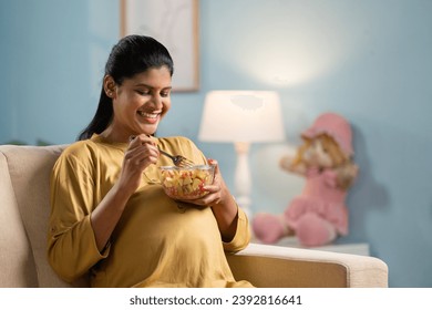 Happy indian pregnant woman eating fruits salad while sitting on sofa at home - concept of healthy lifestyles, Maternal Pregnancy nutrition. - Powered by Shutterstock