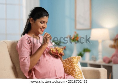 Happy indian pregnant woman eating fruit salad while sitting on sofa at home - concept of Pregnancy nutrition, Healthy eating and Prenatal wellness.