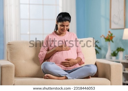 Happy indian pregnant woman feeling baby by looking and holding her tummy at home - concept of motherhood, maternal journey and expecting baby.