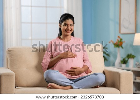 Happy indian pregnant woman feeling baby by looking and holding her tummy at home - concept of motherhood, maternal journey and expecting baby.