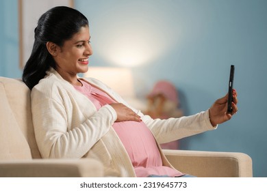 Happy indian pregnant woman making video call on mobile phone while sitting on sofa at home - concept of technology, social media and maternity - Powered by Shutterstock