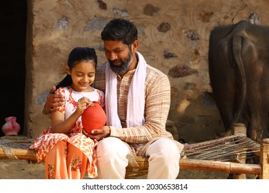 Happy Indian Rural Family Of Father And Daughter Sitting On A Folding Bed In Outdoors Holding A Piggy Bank. Father Teaching His Daughter The Benefits Of Saving Money.