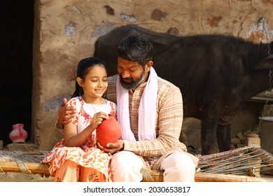 Happy Indian Rural Family Of Father And Daughter Sitting On A Folding Bed In Outdoors Holding A Piggy Bank. Father Teaching His Daughter The Benefits Of Saving Money.