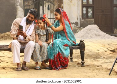 Happy Indian Rural Family Of A Father Mother And A Daughter Sitting Together Engaged With A Mobile Phone. Mother Styling The Hair Of The Daughter. 