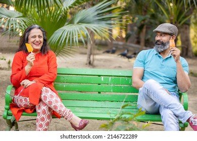 Happy Indian Senior Couple Eating Ice Lolly Or Ice Cream While Sitting On A Bench In A Park Outdoor, Old Mature People Enjoy Retirement Life. Summer Holidays.