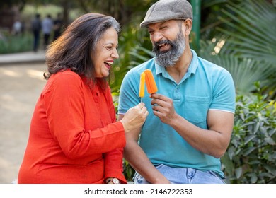 Happy indian senior couple eating ice lolly or ice cream in a park outdoor, old mature people enjoy retirement life. summer holidays.
