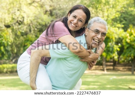 Happy indian senior couple enjoying life having fun at summer park, man giving piggyback ride to woman.