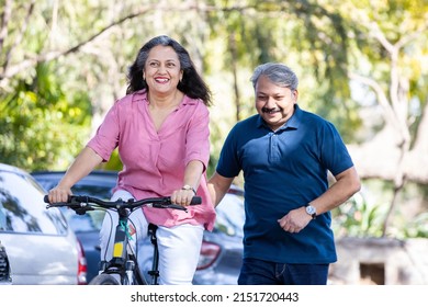 Happy Indian Senior Couple Riding Bicycle In The Park Summer, Active Old Age People And Lifestyle. Elderly Woman Learn To Ride Cycle With Man. Retired People Having Enjoy Life. Selective Focus.