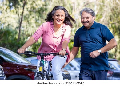 Happy Indian Senior couple riding bicycle in the park summer, active old age people and lifestyle. Elderly woman learn to ride cycle with man. retired people having enjoy life. selective focus.