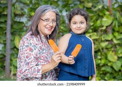 Happy Indian Senior Woman Having Ice Lolly Or Ice Cream With Little Girl Child Together Outdoor, Old Mature People Enjoy Retirement Life. Summer Holidays, Grandmother And Granddaughter Spend Time.