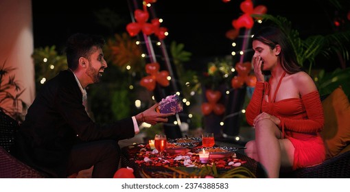 Happy Indian smiling two surprised couple sitting on chair dating outdoor home. Handsome boyfriend gives gift to girlfriend celebrate Valentine's Day spend time enjoying holiday weekend together  - Powered by Shutterstock