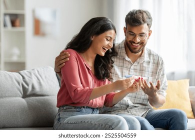 Happy indian spouses holding positive pregnancy test, sitting on couch at home together, couple celebrating awaiting baby, copy space - Powered by Shutterstock