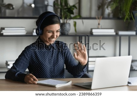 Happy Indian student girl wearing headphones, making video call to teacher, talking to tutor, waving hello at webcam, looking at screen, smiling, writing notes. Woman attending virtual conference chat