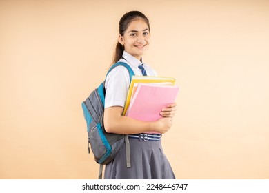 Happy Indian student modern schoolgirl wearing school uniform holding books and bag standing isolated over beige background, Studio shot, Education concept. - Shutterstock ID 2248446747