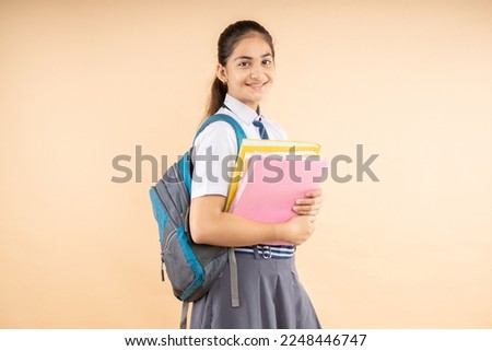 Happy Indian student modern schoolgirl wearing school uniform holding books and bag standing isolated over beige background, Studio shot, Education concept.