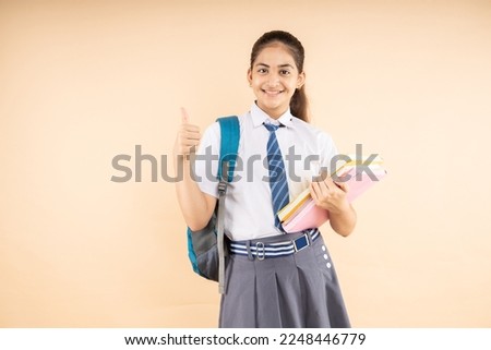 Happy Indian student schoolgirl do thumbs up wearing school uniform holding books and bag standing isolated over beige background, Studio shot, Education concept.