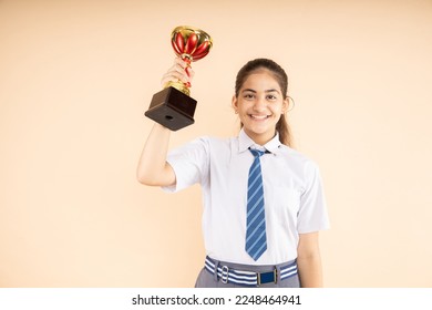 Happy Indian student schoolgirl wearing school uniform holding victory trophy in hand isolated on beige background, Studio shot, closeup, Education concept. - Shutterstock ID 2248464941
