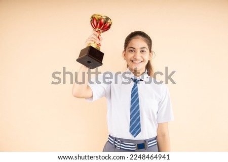 Happy Indian student schoolgirl wearing school uniform holding victory trophy in hand isolated on beige background, Studio shot, closeup, Education concept.