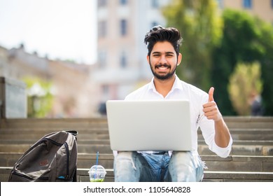 Happy indian student sitting on the stairs showing thumb up working on laptop, in the university campus. - Shutterstock ID 1105640828