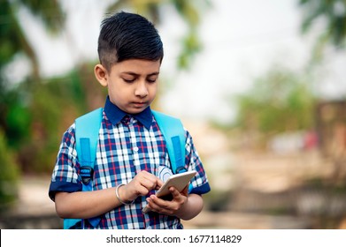 Happy Indian Student Wearing Blue Backpack And Using Smartphone At School Ground. Little School Kid Texting On Mobile Phone While Returning Back To School.