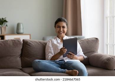 Happy Indian Tablet User Girl Home Portrait. Beautiful Young Millennial Woman Relaxing On Comfortable Sofa, Holding Digital Gadget, Looking At Camera With Toothy Smile. Communication Concept