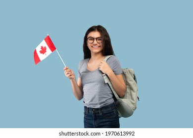 Happy Indian Teen Exchange Student With Backpack Holding Small Flag Of Canada And Smiling At Camera On Blue Studio Background. Travel And Modern Foreign Education Concept